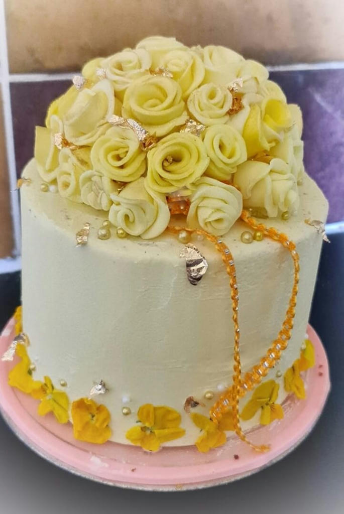 A 6 inch vanilla sponge, homemade jam and buttercream filled. Frosted with pale yellow buttercream and decorated with handmade simple fondant roses, gold leaf, sprinkles and lovely yellow viola